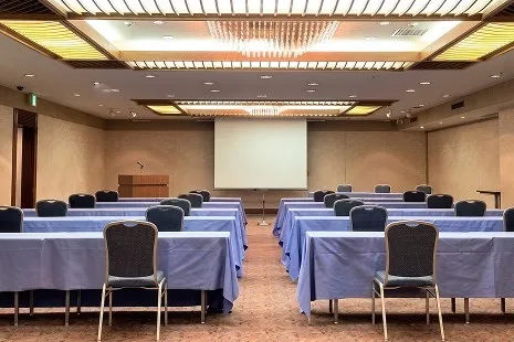 温かみのある木目調の壁に囲まれた、スクール形式（講義スタイル）の落ち着いた中宴会場。前方には大型スクリーンと演台があり、天井中央では繊細なシャンデリア風の照明が輝く上質な空間です。