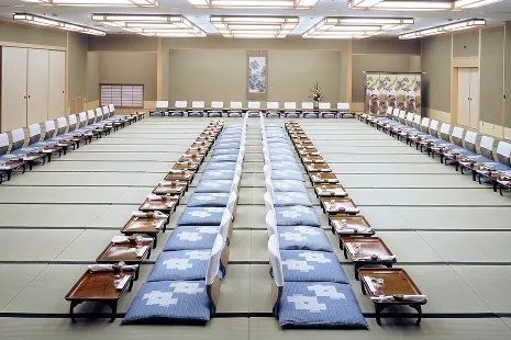 どこまでも広く続く畳敷きの大広間に、お一人ずつの御膳と青い座布団、座椅子が整然と並ぶお座敷スタイルのセッティング。奥の正面には掛け軸が掛けられた床の間や金屏風が飾られた格式高い空間です。