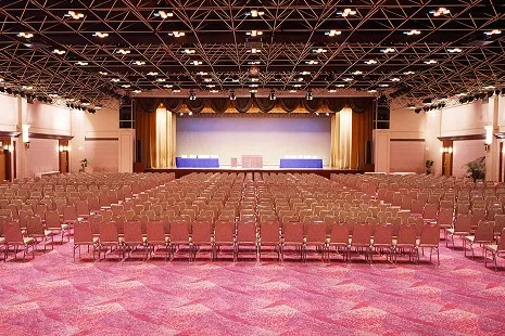 前方にゴールドのドレープカーテンが飾られた本舞台（ステージ）がある、シアター形式（講演会仕様）の大宴会場。ステージに向かって椅子が整然と敷き詰められており、天井はむき出しのトラス構造に無数のスポットライトが設置された広大な空間です。
