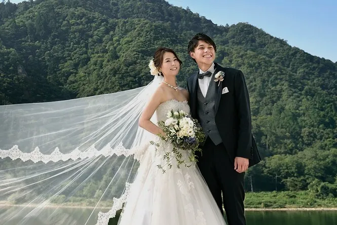 青々と茂る雄大な金華山と、穏やかに流れる長良川をバックに微笑み合う新郎新婦のウエディングポートレート。新婦の美しいレースのロングベールが風になびいて大きく広がる、ドラマチックで開放感あふれるカットです。