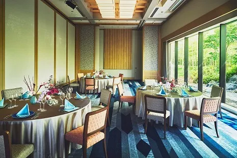 右側の大きな窓から差し込む光と、外に見える竹林の緑が美しい中宴会場。室内には、クロスが掛けられた丸テーブルが配置され、青いナプキンやワイングラス、装花が華やかにセットされた宴席仕様の空間です。