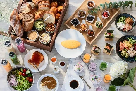 光が差し込むテーブルに並ぶ、彩り豊かな朝食ビュッフェの全景。バゲットやクロワッサンが詰まったカゴ、中央の白い皿にのった綺麗なオムレツ、新鮮なグリーンサラダ、ご飯にのったカレー、スモークサーモンなどの冷菜、数種類の小鉢、ミニサイズのり巻き、カラフルなボトルドリンクやカップスープが美しくディスプレイされています。