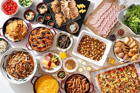 真上から見下ろした、大皿料理がひしめき合う豪華なビュッフェテーブル。大きな四角いピザ、山盛りのソーセージ、スクランブルエッグ、具だくさんの筑前煮、トマトパスタ、麻婆豆腐のような炒め物、きれいに並んだ焼き魚や大量のハム、カゴに入ったパン、新鮮なサラダやミニトマトなどが画面いっぱいに広がっています。