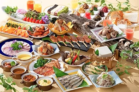 テーブルいっぱいに並ぶ、岐阜の特産を活かした和洋バイキングの料理。中央に並んだ串付きの五平餅、手前の若鮎の天ぷらや焼き魚、ベーコンやスクランブルエッグ、肉じゃが、冷奴、さらに原木椎茸を模したディスプレイや、色鮮やかな大皿サラダ、カットフルーツ、一口ケーキなどが華やかに並んでいます。