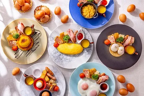 真上から見下ろした、さまざまな卵料理が主役の洋朝食プレート。中央のプレーンオムレツをはじめ、エッグベネディクト、目玉焼き、スクランブルエッグ、ゆで卵、フレンチトースト風の仕上がりなど、それぞれ異なる卵料理が色とりどりの丸皿に美しく盛られ、まわりにはたくさんの生卵が散りばめられています。