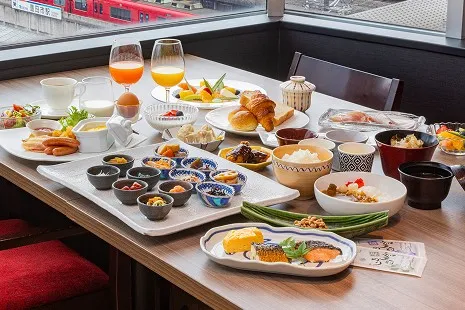 窓際のテーブル席に並ぶ、彩り豊かな和洋朝食の全景。明太子やひじき煮など、一口サイズのおかずが入った多数の小さな小鉢を中心に、焼き魚、ご飯と味噌汁、焼き立てのパンやソーセージ、サラダ、フルーツ、グラスに注がれたジュースなどが上品にディスプレイされています。