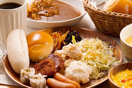 木の丸皿に賑やかに盛り付けられた朝食バイキングの一皿。左側には白いコッペパンと丸いロールパン、手前にはシュウマイ2個、ウインナー、カツが並び、右側には山盛りの千切りキャベツ、ポテトサラダ、蒸し鶏、奥にはきんぴらごぼうやひじき煮がのっています。背景にはカレーライスやコーヒーも添えられています。