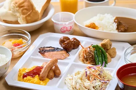 テーブルに並ぶ和洋朝食ビュッフェの料理。手前の白い仕切り皿には、ケチャップのかかったスクランブルエッグとソーセージ、焼き魚、ミートボール、煮物やインゲン、千切りキャベツのサラダが盛られています。その後ろにはカレーライス、カゴに入った食パン、カップに入った納豆、お味噌汁、ジュースやコーヒーが並んでいます。