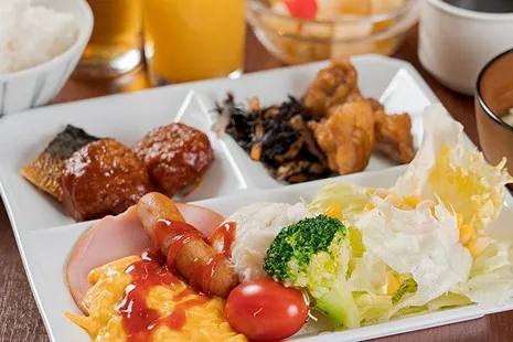 スクランブルエッグやソーセージ、新鮮なサラダといった洋食メニューと、焼き魚、ひじきの煮物、唐揚げなどの和食惣菜が、四角いプレートに彩りよく少しずつ盛り付けられた朝食ビュッフェのおかずの一皿。