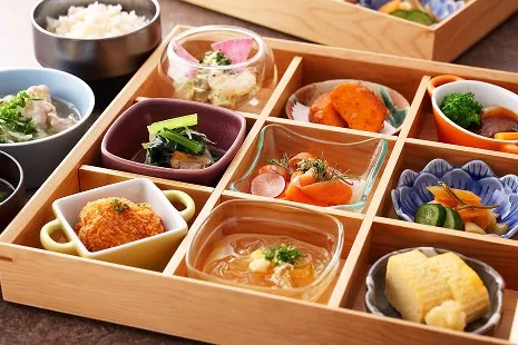 上品な木製の9マス仕切り箱に、色とりどりの小鉢が一つずつ美しく並んだ朝食ビュッフェの一膳。ガラスの器に盛られたスモークサーモンやところてん、エビの和え物をはじめ、厚焼き玉子、お漬物、おひたし、一口サイズのカツなどがそれぞれ異なる形の小さな器に少しずつ盛り付けられています。左奥には白ご飯や、お肉の小鉢も添えられています。