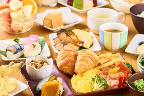 テーブルいっぱいに並ぶ、品数豊富な和洋朝食ビュッフェの料理。手前の紫の仕切り皿には、焼き立てのクロワッサン、スクランブルエッグ、フライドポテト、お肉の炒め物などが盛られています。その周りには、サクサクとした天ぷら、色鮮やかな手まり寿司、大ぶりの焼き魚、カットフルーツや一口ケーキ、ご飯とお味噌汁などが贅沢に並んでいます。