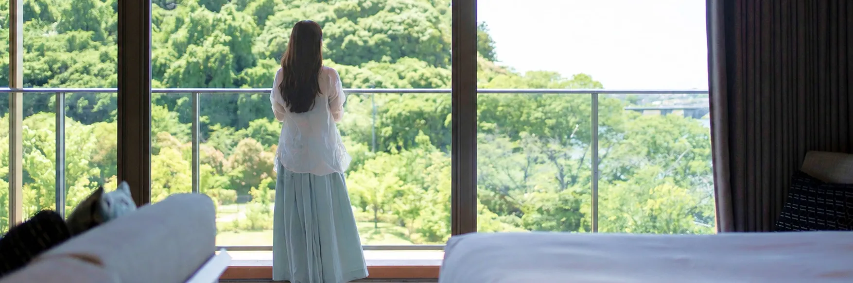 緑豊かな木々を見渡せる広々とした客室のテラスで、外の景色を眺めながら佇む女性の背中。