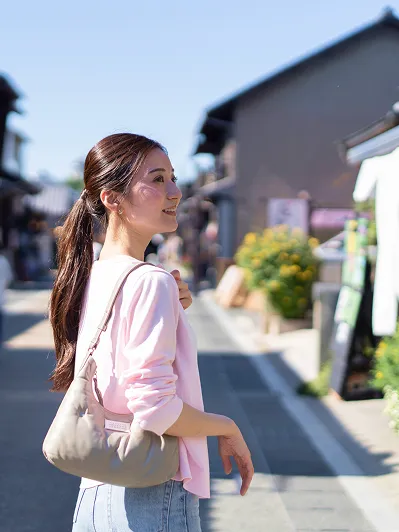 晴天のもと、和風の古い街並みをバックに、肩にバッグをかけて笑顔で歩く旅行中の女性。