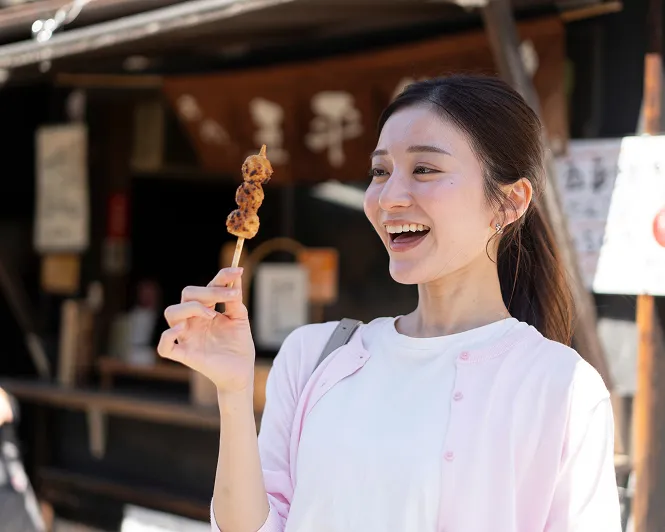 和風の古いお茶屋をバックに、串に刺さったお団子を手に嬉しそうに微笑む旅行中の女性。
