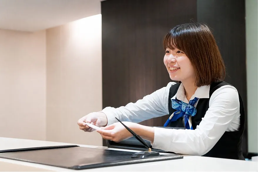 清潔感のあるフロントカウンターで、にこやかな笑顔で優しく宿泊客を迎える名鉄インの女性スタッフ。