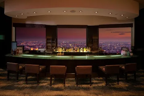 「バー スターゲイト」のカウンター席。大きな窓の向こうに美しい夕焼けと広大な街並みが広がり、カウンターにはお酒のボトルが並んでいます。