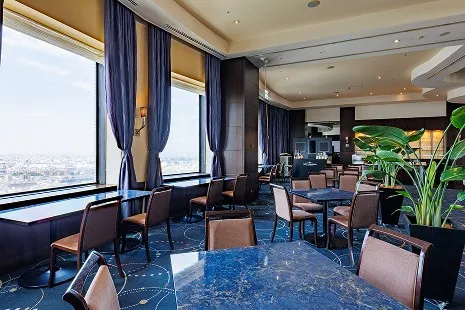 「ウイングラウンジ」の内観。大きな窓から明るい青空と街並みが一望できる店内に、落ち着いた色合いのテーブル席が並んでいます。